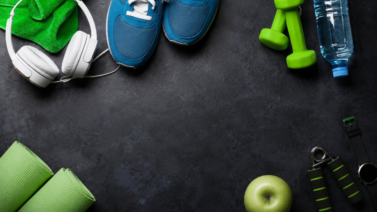 A styled flat-lay of fitness and gym accessories on a dark textured surface, including blue athletic shoes, white over-ear headphones, green dumbbells, a water bottle, rolled green exercise mats, a green apple, jump rope handles, and a green towel.