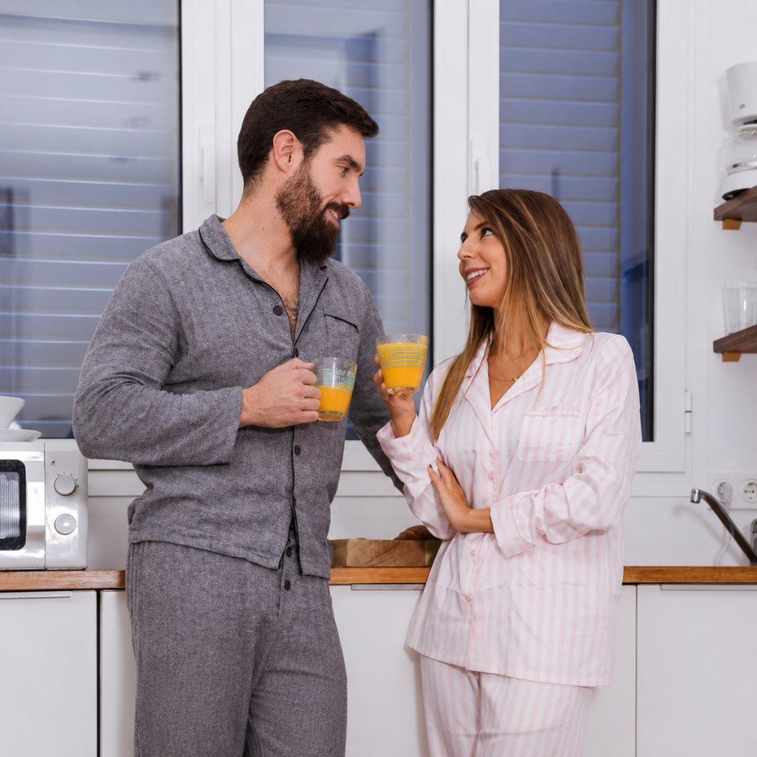 A man and a woman in matching pajamas stand in a bright kitchen, smiling at each other while holding glasses of orange juice. The man wears a gray long-sleeve pajama set, and the woman wears a light pink and white vertically striped pajama set.
