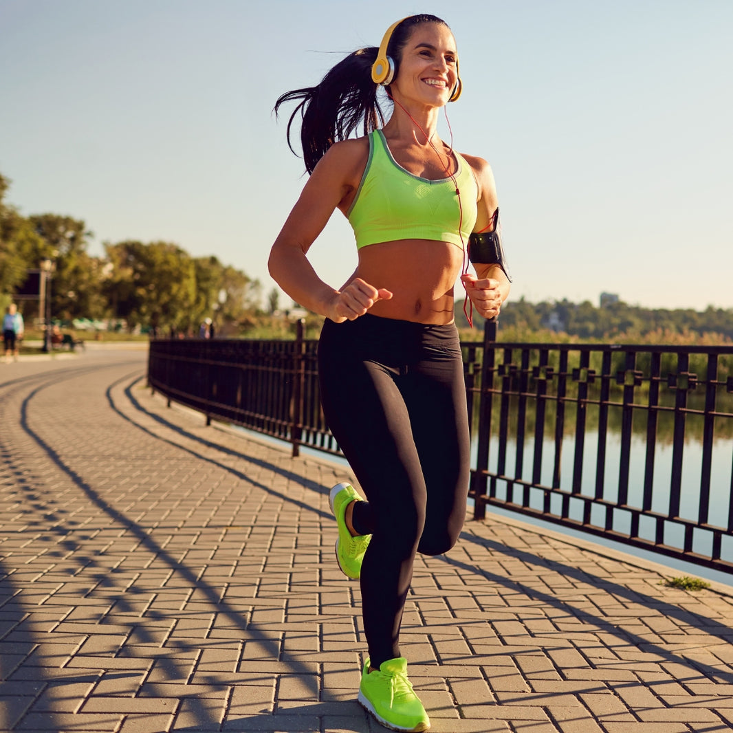 A woman jogging outdoors on a paved path beside water, wearing a bright neon green sports bra, black leggings, neon green running shoes, and yellow headphones, with a black armband holding a phone.