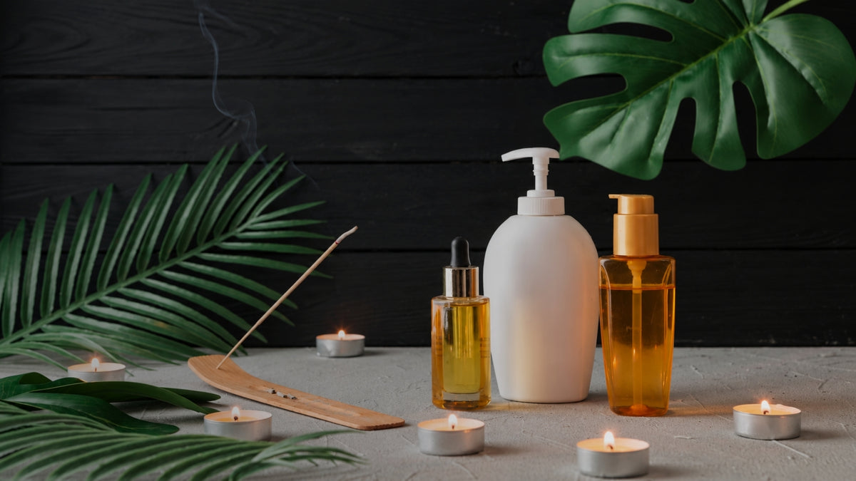 A spa product set featuring a white pump bottle, an amber glass dropper bottle, and an amber glass bottle with a gold cap, styled with green tropical leaves, an incense stick, and burning tea lights on a dark wood background.