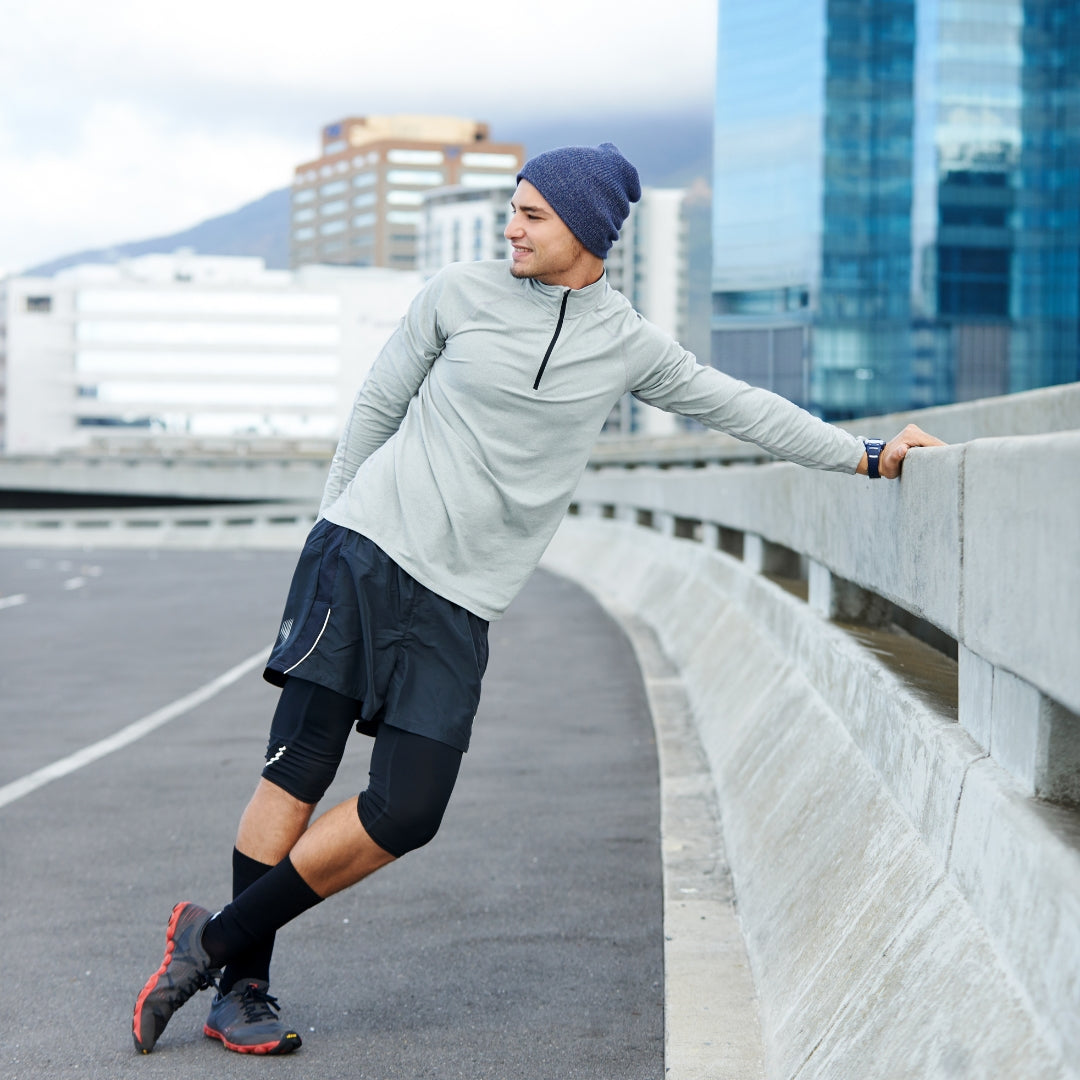 A man in athletic wear, including a light gray long-sleeve quarter-zip top, black shorts, black compression sleeves, and a navy beanie, leans against a concrete bridge railing in an outdoor urban setting with skyscrapers in the background.